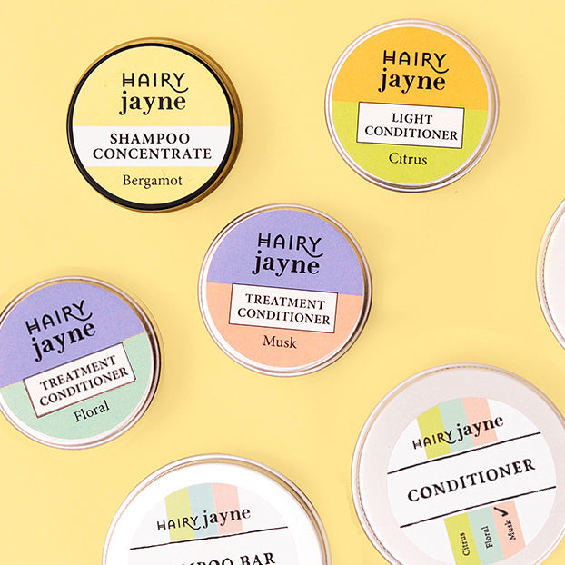 Six round haircare product tins arranged on a pale yellow background. The visible brands and products include Hairy Jayne Shampoo Concentrate in Bergamot, Hairy Jayne Light Conditioner in Citrus, Hairy Jayne Treatment Conditioner in Musk, Hairy Jayne Conditioner, and two other Hairy Jayne items.