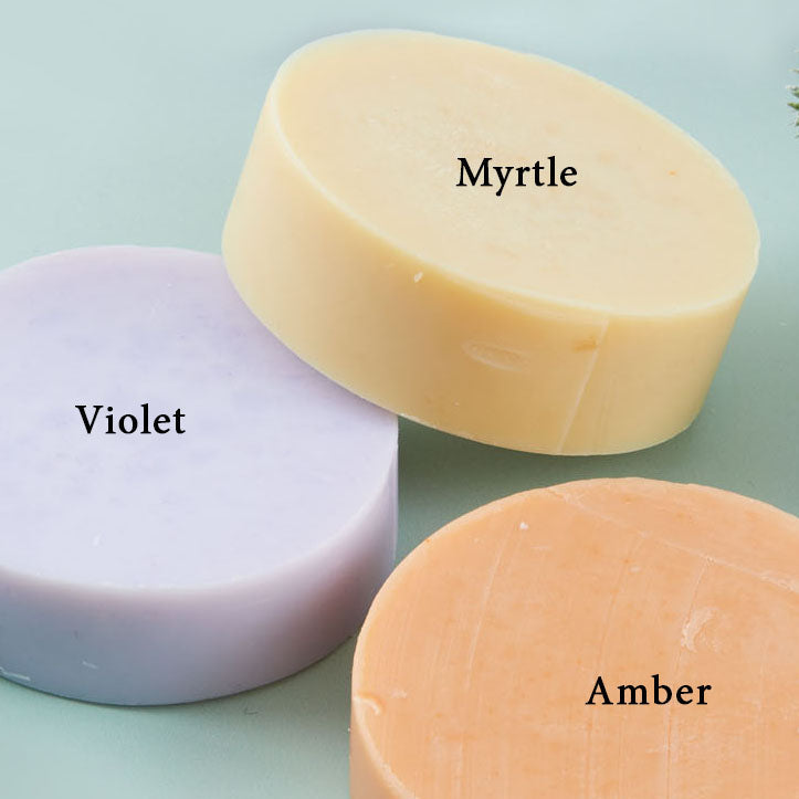 Three different colored soaps labeled 'Violet', 'Myrtle', and 'Amber' on a light blue background with greenery.