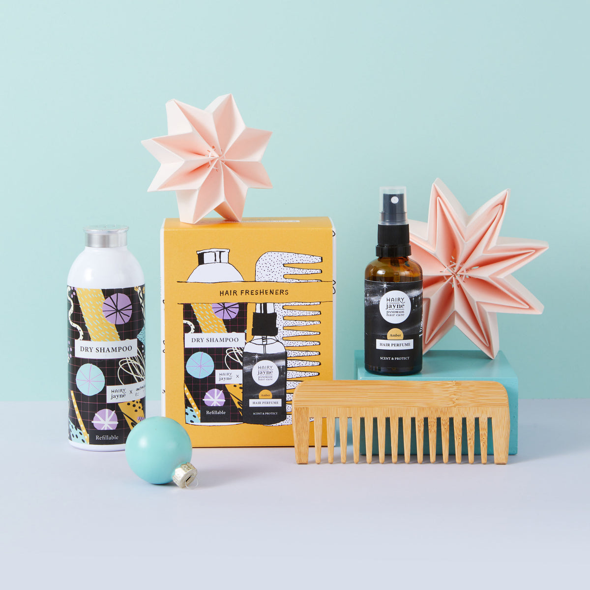 A hair care gift set arranged on a pale blue and white surface against a light aqua background. The set includes a bottle of dry shampoo, a boxed hair freshener, a small amber spray bottle, a wooden comb, and two pink origami stars.