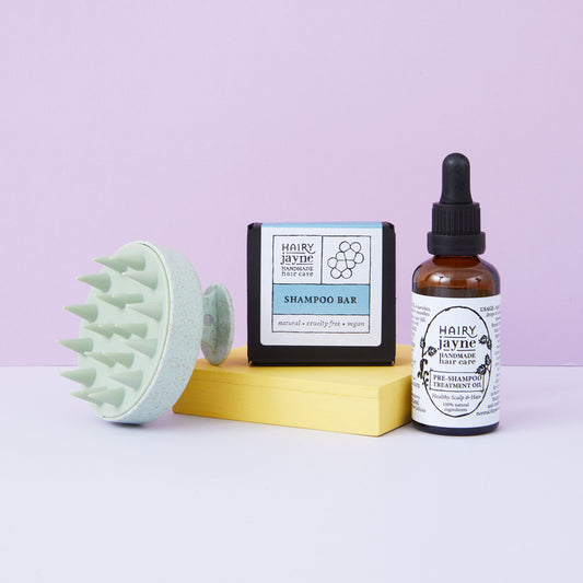 Hairy Jayne hair care products including a scalp massager, shampoo bar, and oil on a light purple background.