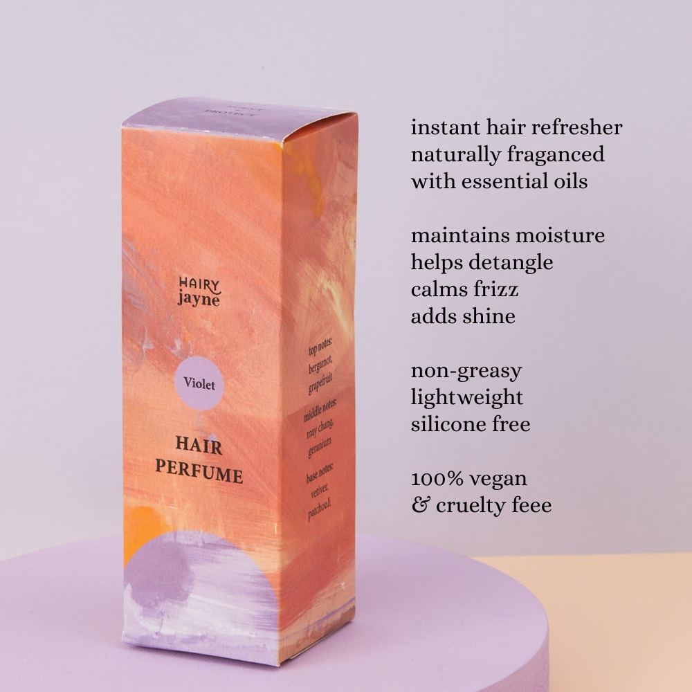 A box of Hairy Jayne Hair Perfume in the Violet scent, displayed on a lavender pedestal with descriptive text on the right against a light lavender background.