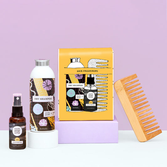 A hair care gift set featuring a yellow box of hair fresheners, a white bottle of dry shampoo, a small amber spray bottle of hair oil, and a wooden wide-tooth comb, arranged on a white surface with a lavender background.