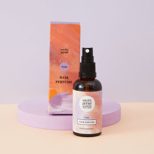 An amber glass spray bottle of Hairy Jayne Violet Hair Perfume stands in front of its orange and lavender packaging box on a pastel lavender and peach studio set.