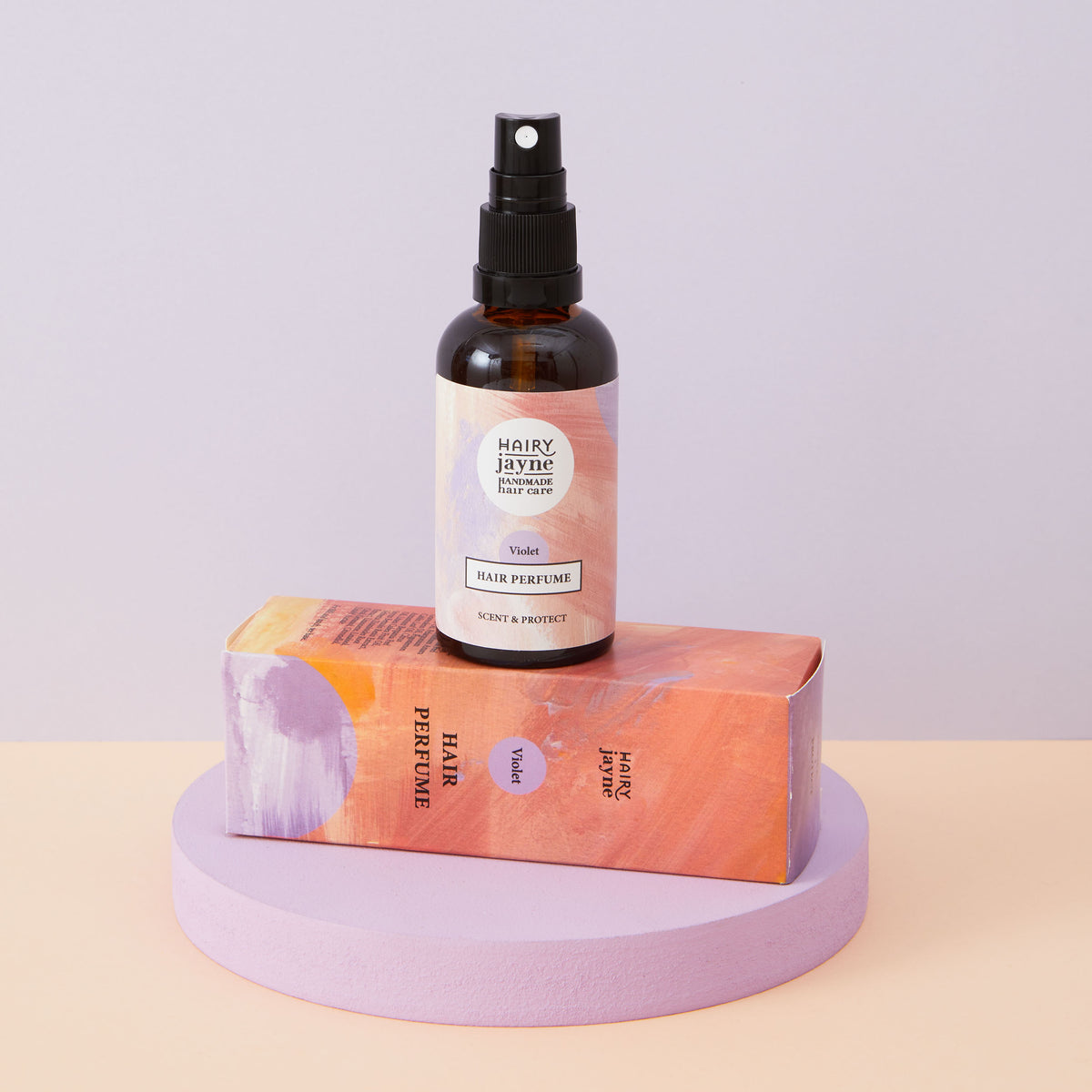 An amber glass spray bottle stands upright on top of its matching peach and lavender box, set on a round lavender pedestal against a pale lavender background.