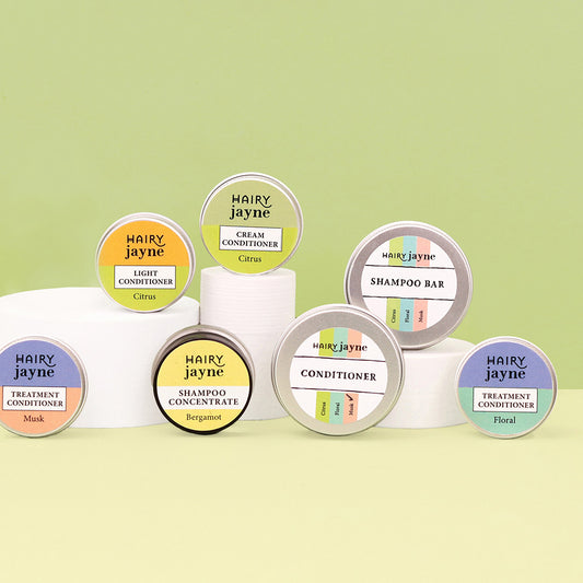 A set of Hairy Jayne haircare containers arranged on white cylindrical stands against a pale green background. Visible products include a mask, light conditioner, cream conditioner, shampoo bar, conditioner, shampoo concentrate, and two treatment conditioners, each with round lids showing different pastel color labels.