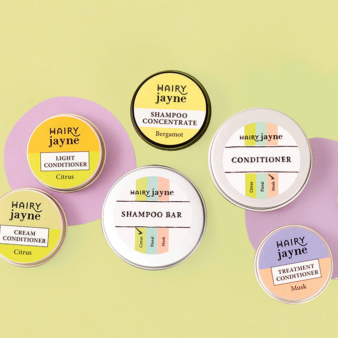 Six round cosmetic tins from Hairy Jayne are arranged on pastel lavender and yellow circular backdrops against a pale yellow-green background. The products include Shampoo Concentrate (Bergamot), Light Conditioner (Citrus), Cream Conditioner (Citrus), Shampoo Bar, Conditioner, and Treatment Conditioner (Musk).