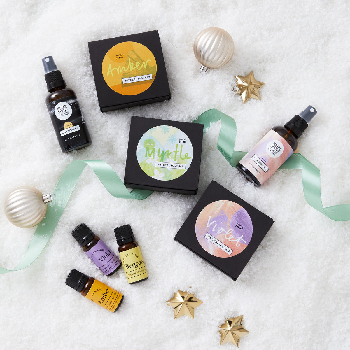 A flat-lay arrangement of essential oils and aromatherapy products on a white textured surface with decorative gold stars and a mint ribbon. Visible items include black boxes labeled Amber, Myrtle, and Violet, along with two amber spray bottles and three small amber essential oil bottles with purple, yellow, and green labels.