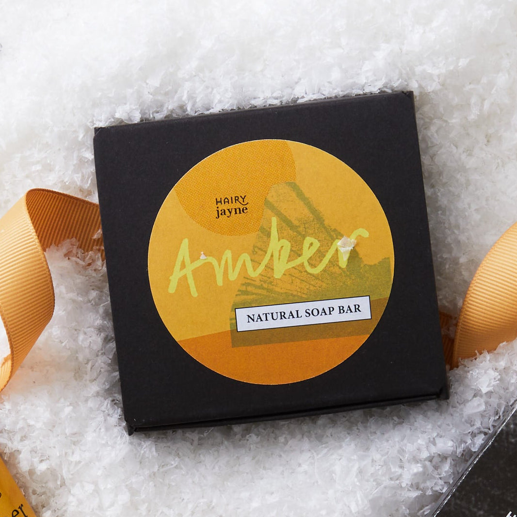 A styled flat-lay of amber-themed hair care products on a white textured background resembling snow. The set includes a black square box labeled 'Amber Natural Soap Bar', a small amber bottle with a yellow label, a black rectangular box labeled 'Hair Perfume', and decorative gold ribbons and metallic ornaments.