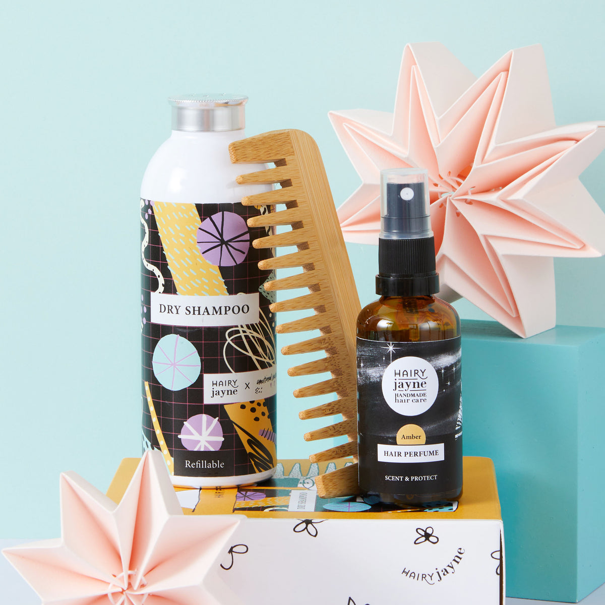 Haircare gift set from Hairy Jayne featuring a white dry shampoo bottle with a colourful patterned label, a wooden hair comb, and a small amber spray bottle of hair perfume, styled on a yellow box with pink origami stars against a light blue background.
