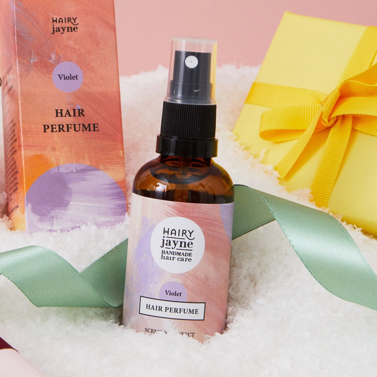 A bottle of Hairy Jayne hair perfume and its packaging box are displayed on white faux snow alongside gift boxes, a green ribbon, and a gold ornament against a solid pink background.