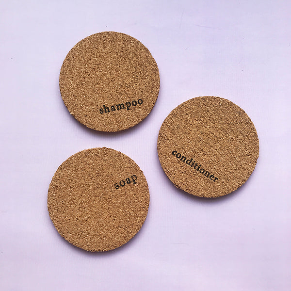 3 circular mats made from cork, they are stamped with the words shampoo, conditioner and soap